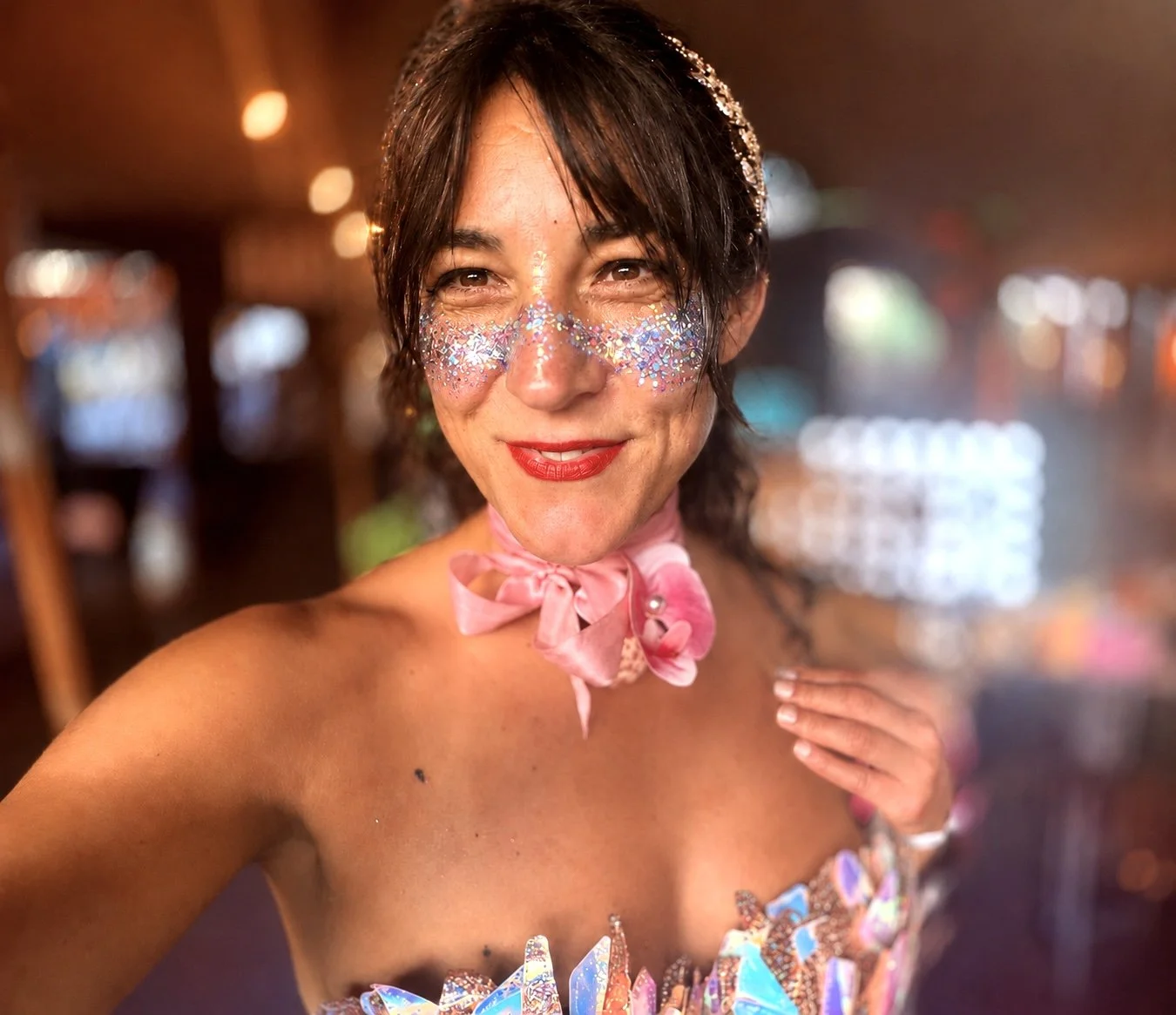 A woman with glitter and face paint, smiling, in colorful festival attire with a pink ribbon around her neck, in a lively environment.