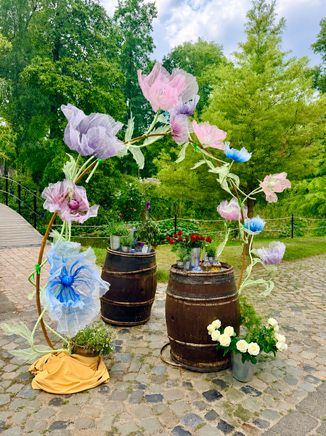 Decorative floral display with large colorful fabric flowers on arched supports, set on cobblestone ground in a garden, with barrels used as plant stands and lush green trees in the background.
