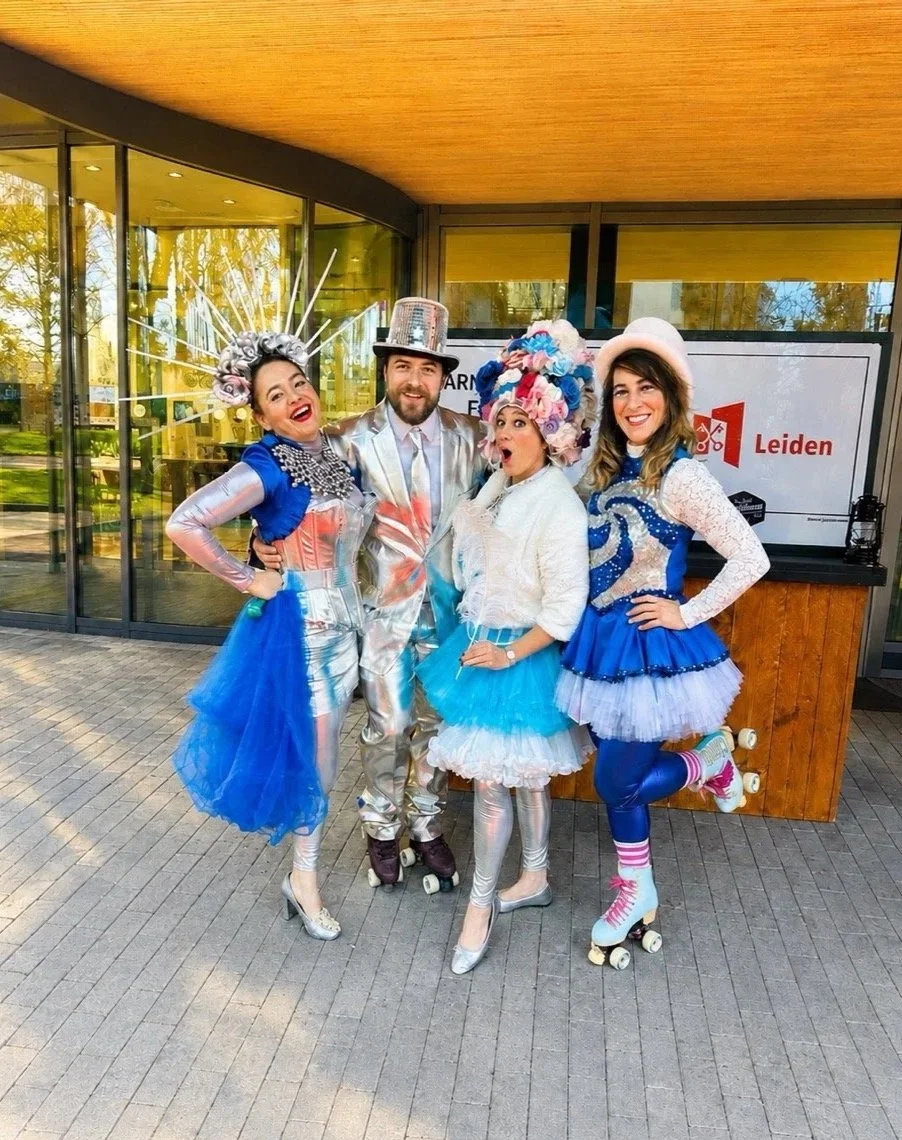 Four people dressed in colorful and whimsical costumes, including tutus, shiny tights, and roller skates, posing outdoors in front of a building with glass windows and a sign that says 'Leiden'.