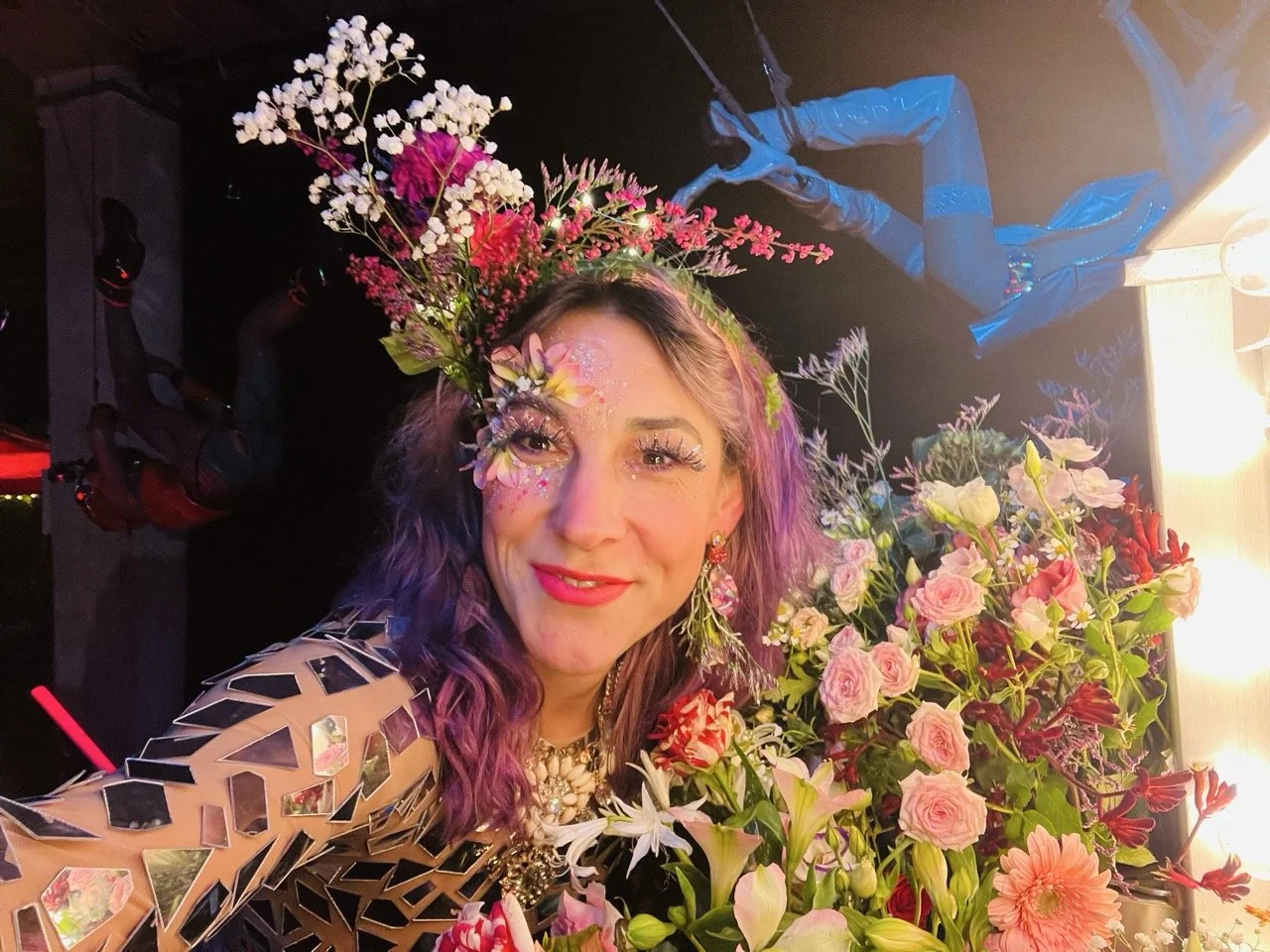 A woman with purple hair and floral makeup, wearing a metallic geometric dress, posed next to a large bouquet of pink and white roses and other flowers, in a colorful setting with decorative lighting and blue duct tape in the background.