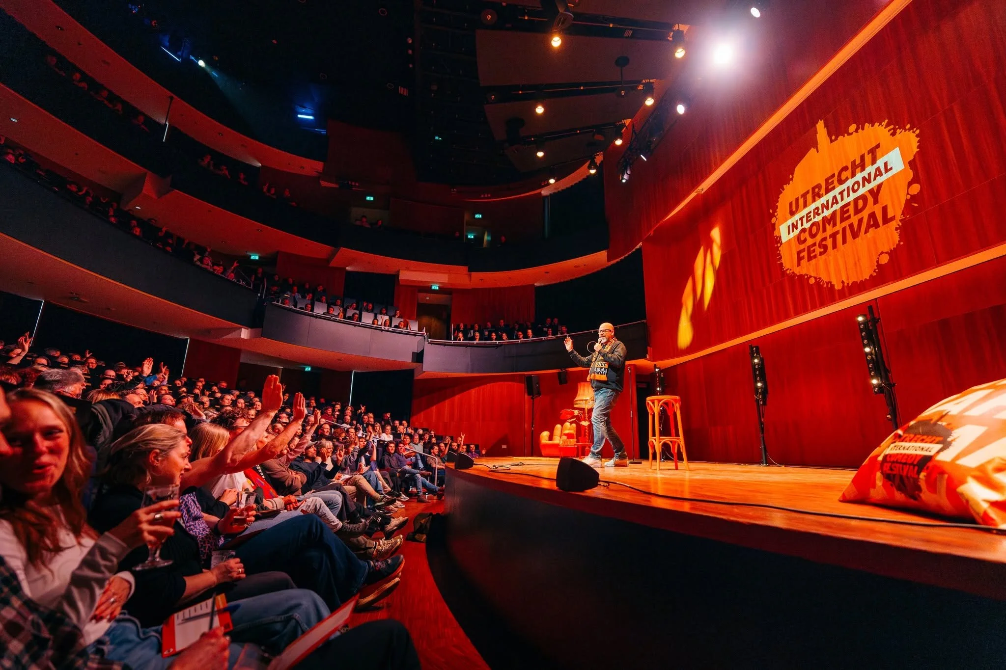 Audience attending the Utrecht International Comedy Festival, with a performer on stage in a theater with red walls and curtains, and a large orange sign with event name.