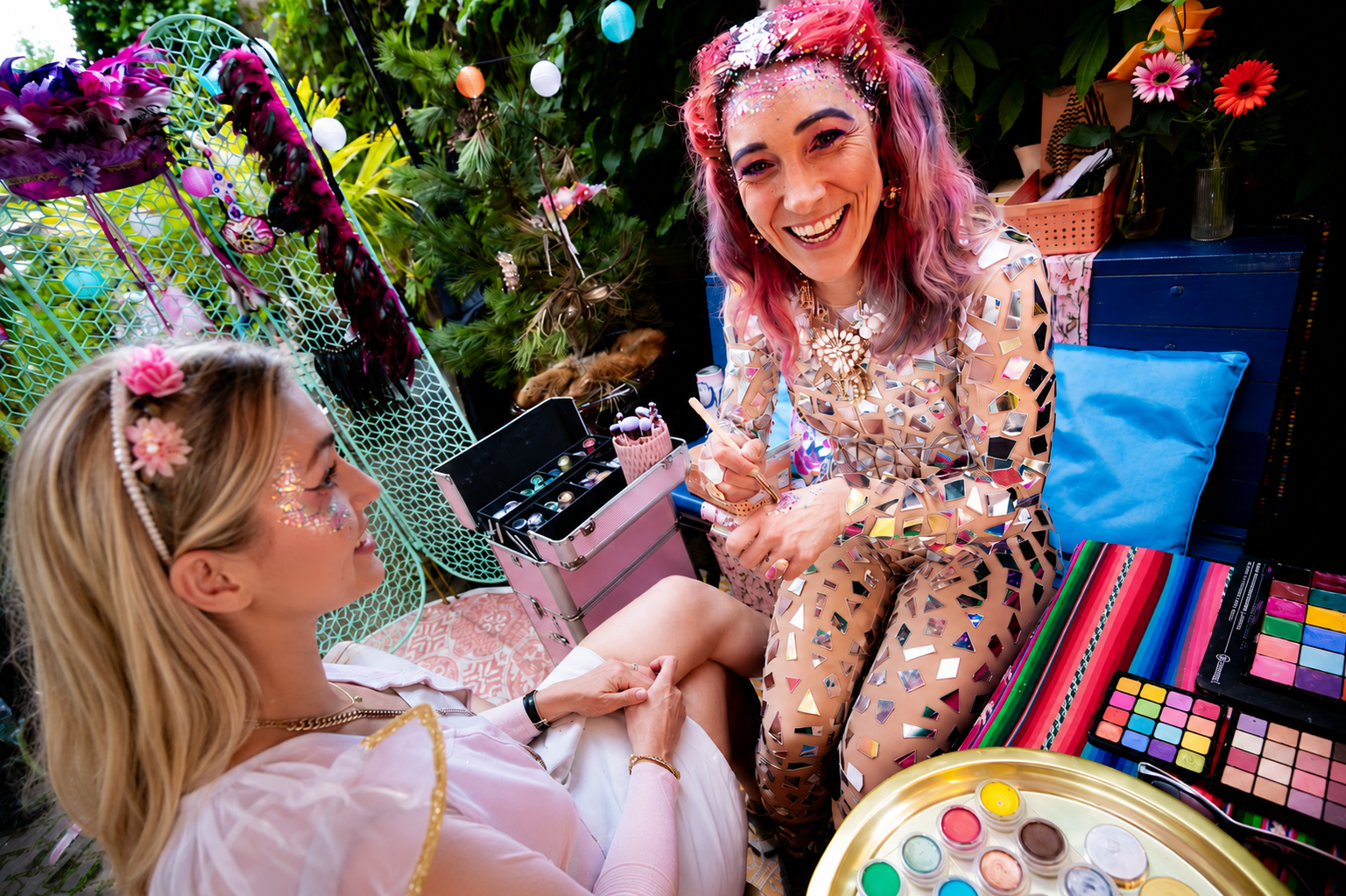 Two women at a colorful makeup and accessories station, one sitting and the other leaning down, smiling. The woman sitting has face glitter and flowers in her hair, the other woman has pink hair and a mirror mosaic outfit, surrounded by makeup palettes, jewelry, and decorative flowers.