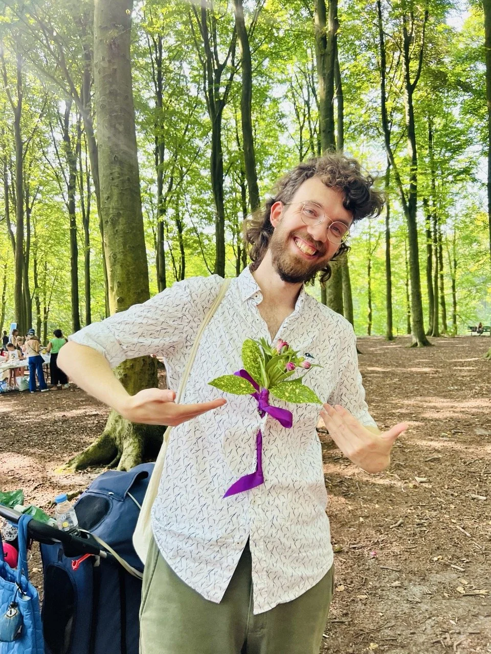 A man with glasses and curly hair standing outdoors in a forest, smiling and pointing to a flower arrangement on his chest made of green leaves, purple ribbon, and small flowers.