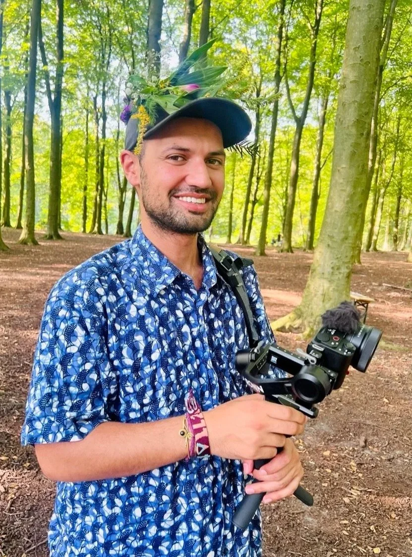 A man smiling in a forest, wearing a blue patterned shirt, a hat decorated with flowers and leaves, and holding a camera stabilizer.