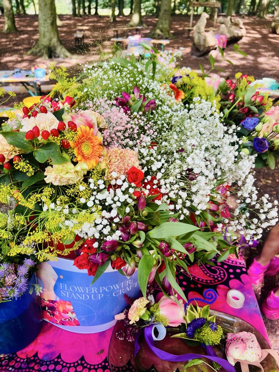 A colorful bouquet of various flowers including baby's breath, roses, and others, placed on a pink table with a sign that reads 'Flower Power: Stand Up + Craft', outdoors in a wooded area.