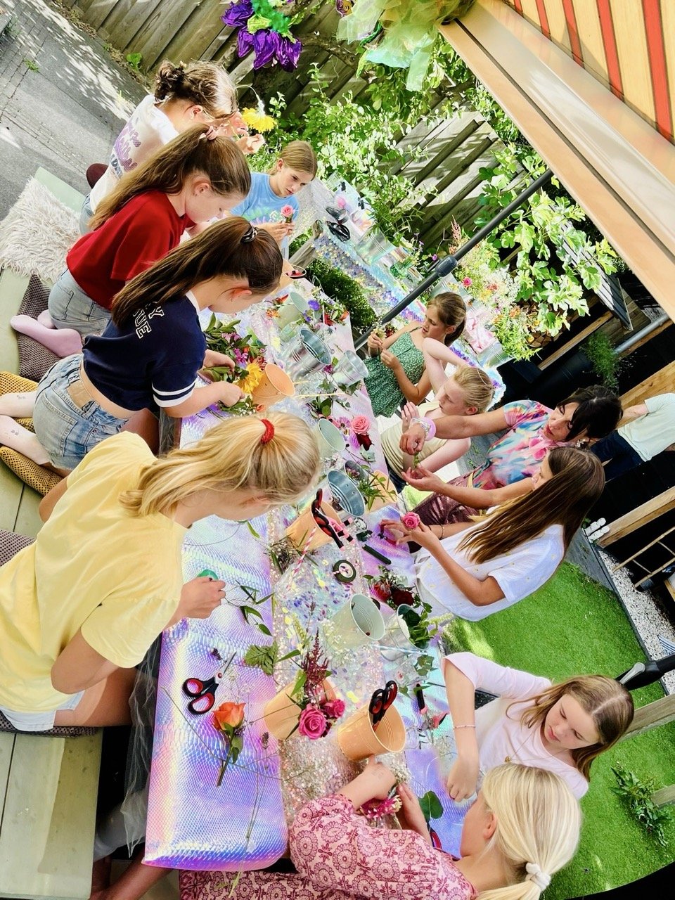 Children participating in a flower arrangement activity outdoors at a long table covered with a holographic tablecloth, surrounded by green plants and flowers, with an adult assisting them.
