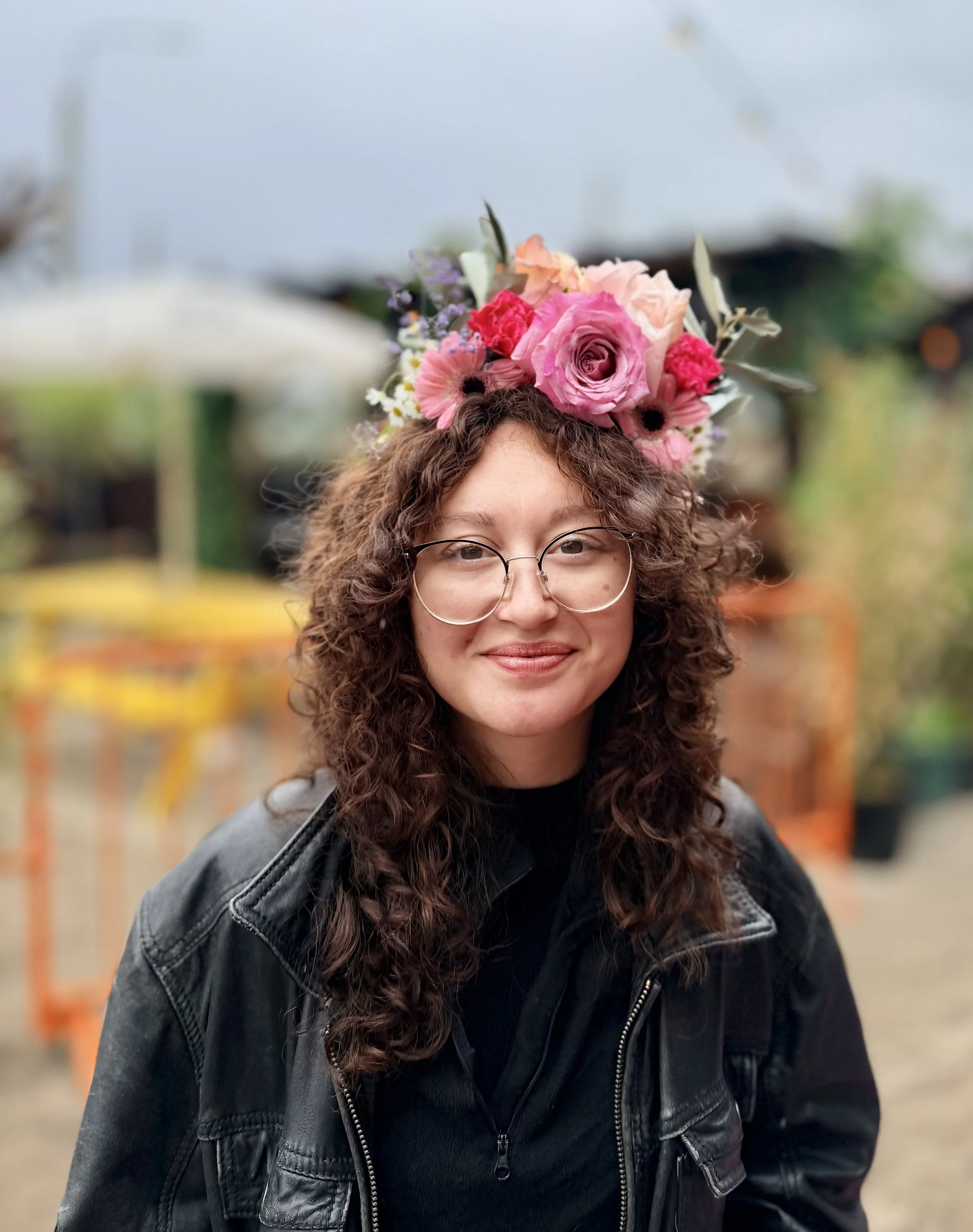 A person with curly hair wearing glasses and a black jacket, adorned with a large colorful flower crown, smiling in an outdoor setting.