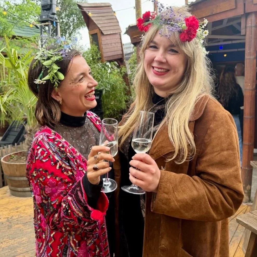 Two women wearing floral crowns and holding champagne glasses, smiling at each other at an outdoor gathering.