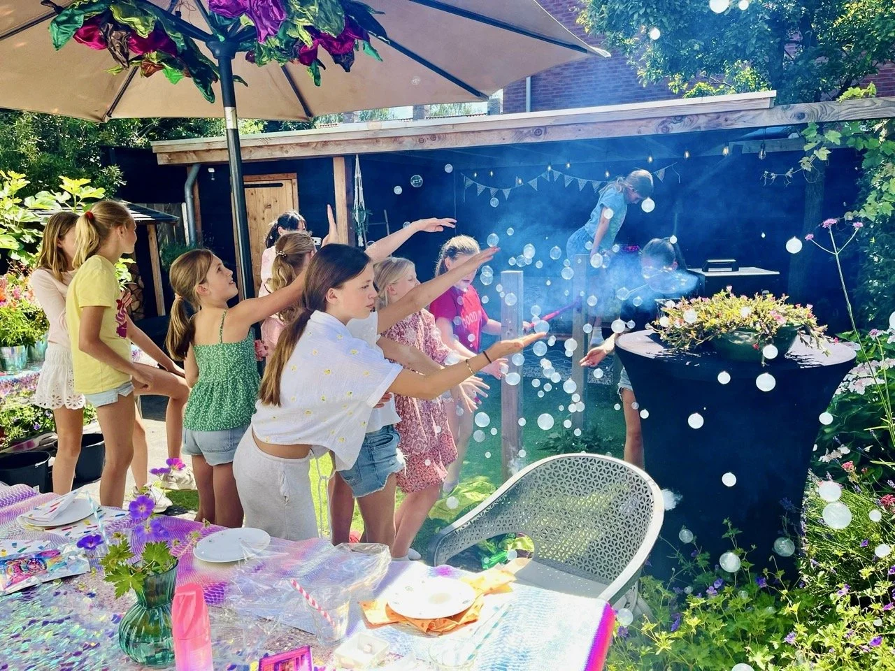 Children at a birthday party outdoors, with some reaching out towards a bubble machine, under a large umbrella with decorated tables and a garden backdrop.