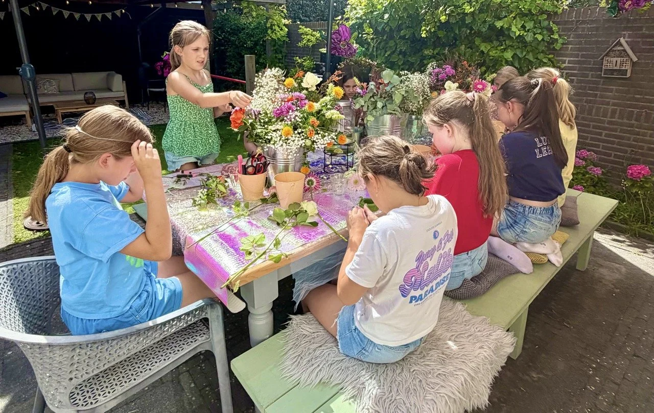 Children participating in a flower arranging activity outdoors around a table filled with flowers, scissors, and gardening supplies.