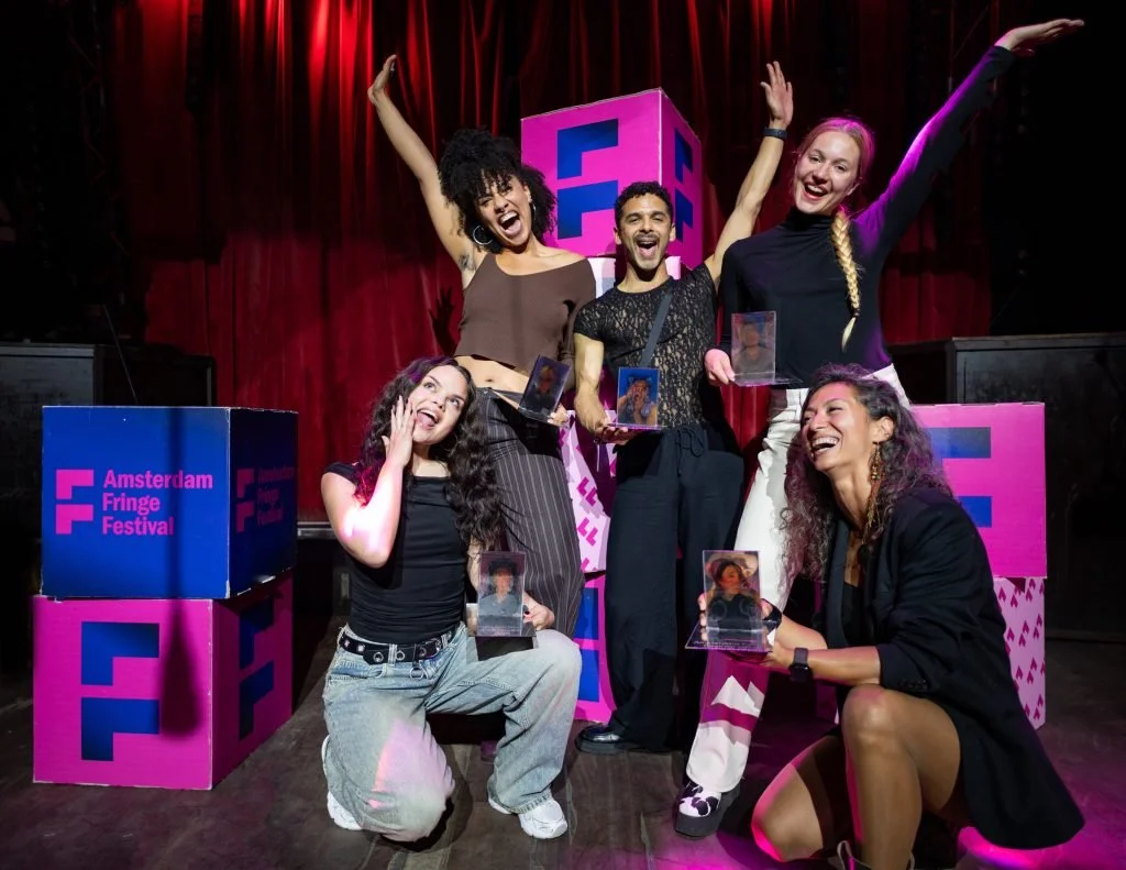 Group of five diverse people celebrating on stage at the Amsterdam Fringe Festival, holding awards, with red curtains and festival branding in the background.