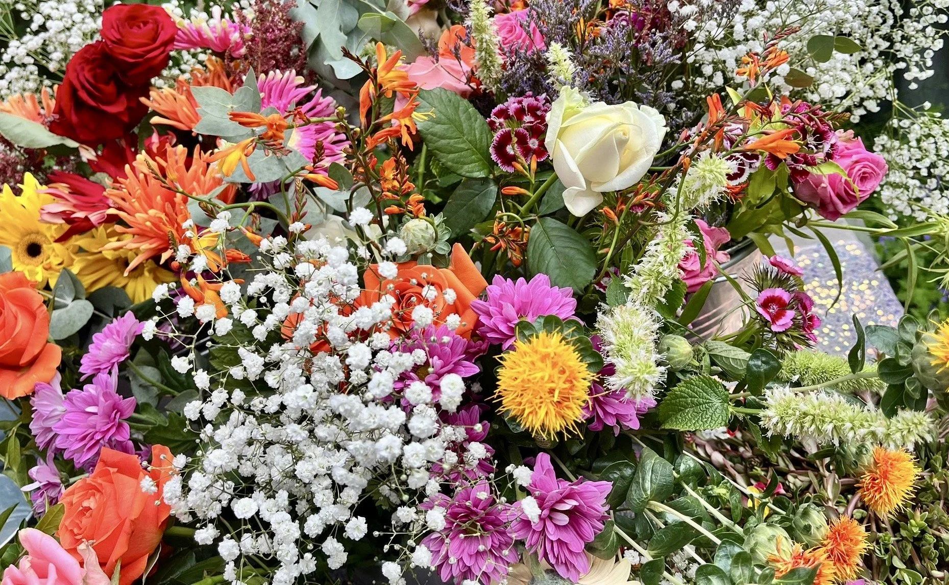 A colorful assortment of fresh flowers, including roses, daisies, baby's breath, and other blossoms in shades of red, pink, orange, yellow, white, and purple, with green leaves and foliage.