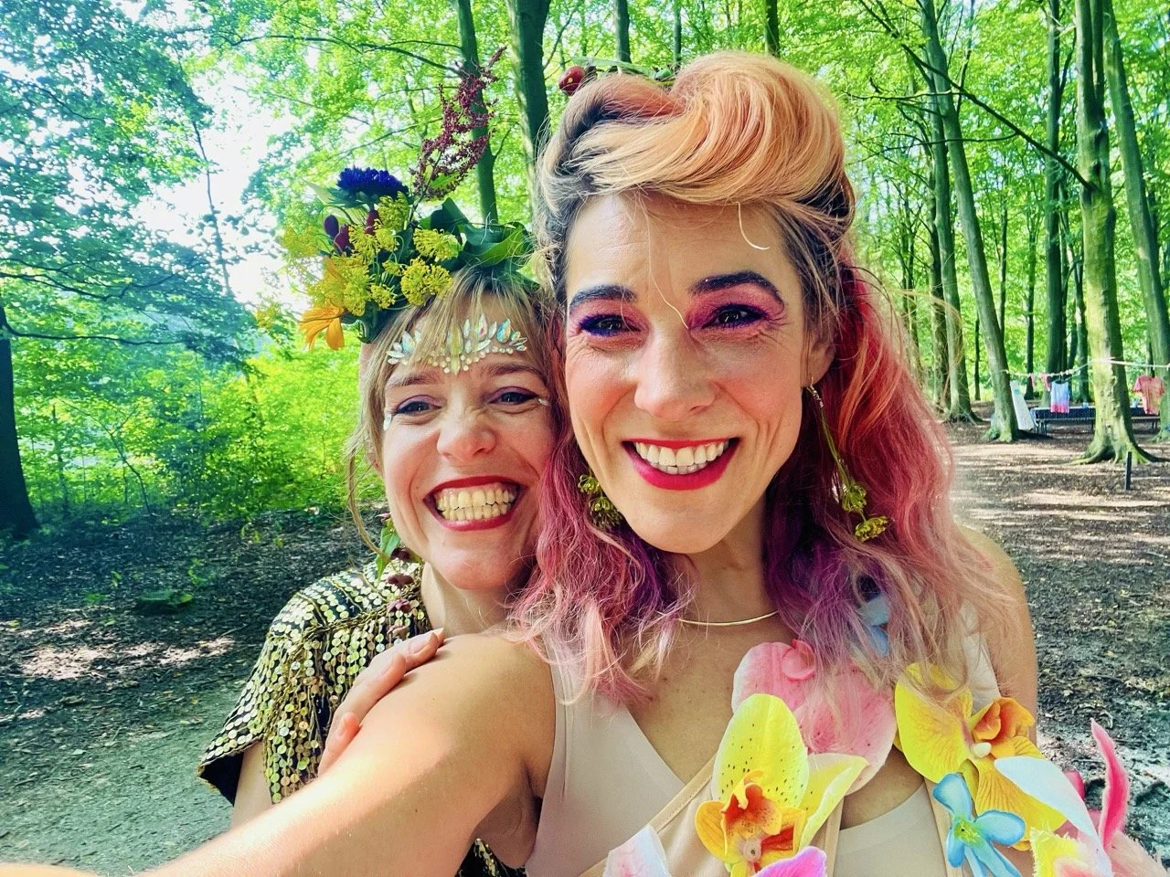 Two women smiling in a forest, one with pink and purple hair wearing a white dress and floral jewelry, the other with blonde hair, a floral crown, and wearing a sequined top.