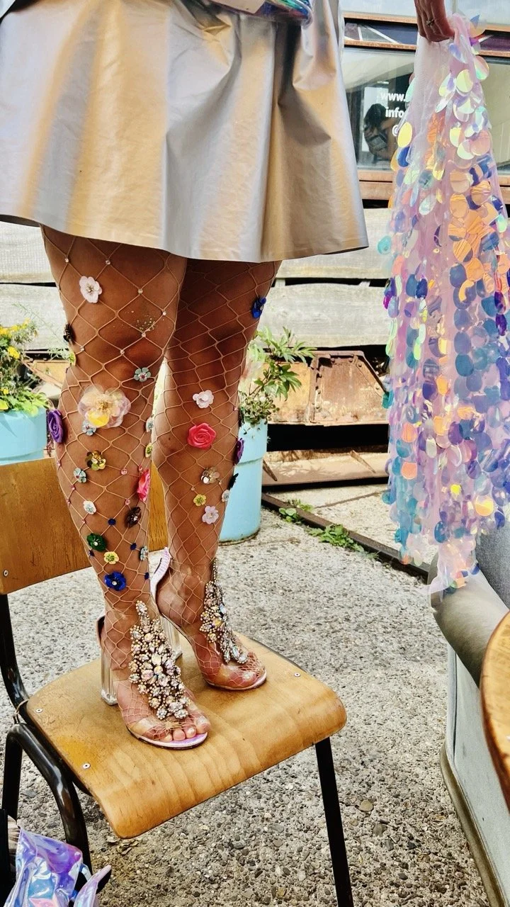 Person standing on a wooden chair wearing a beige skirt, glittery high heels decorated with large floral embellishments, fishnet stockings adorned with colorful flower patches, and holding a shiny, iridescent dress.