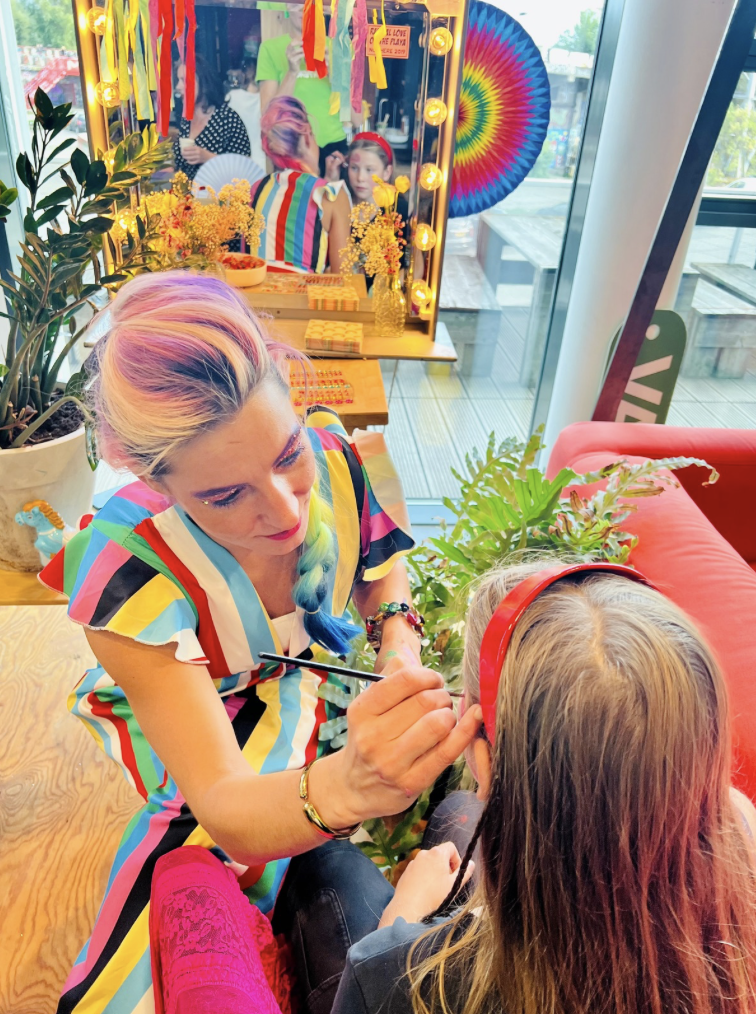 A woman with colorful hair and a rainbow-striped dress is applying face paint to a young girl's face, who is wearing a red headband and black shirt, in a vibrant, decorated room with plants, colorful ribbons, and a tie-dye backdrop.