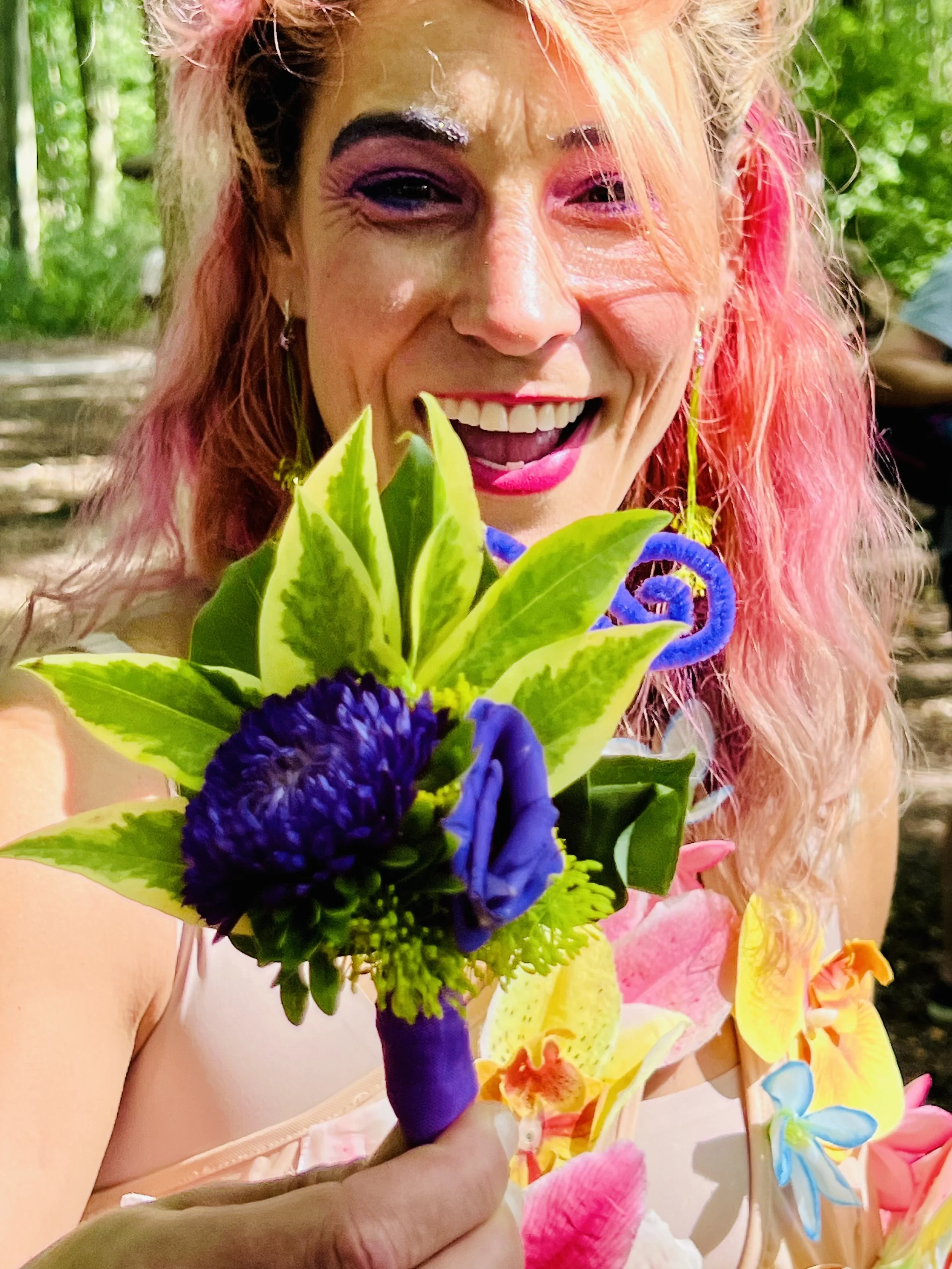A woman with colorful, vibrant makeup and pink hair holding a bouquet of flowers, dressed in a colorful, floral party dress, smiling outdoors.