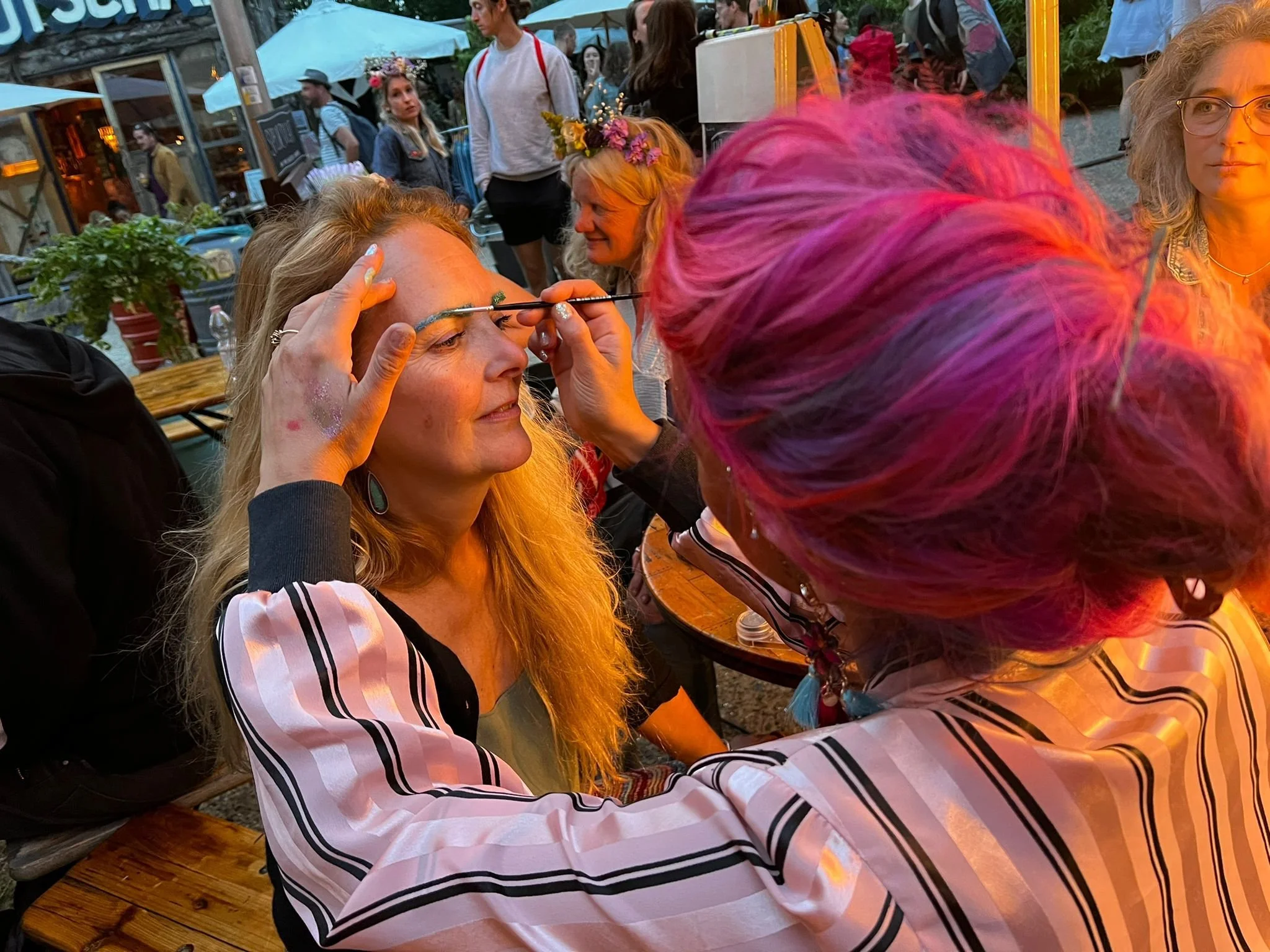 A woman with long blonde hair and colorful face makeup sitting at a table, another woman with bright pink hair leaning in, and other people in the background at an outdoor event or festival.