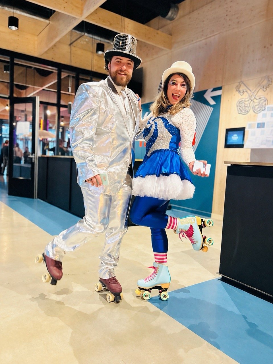 Two people dressed in roller skating costumes, one in a shiny silver suit with a tall top hat and the other in blue and white with a cream hat, posing and smiling while skating indoors.