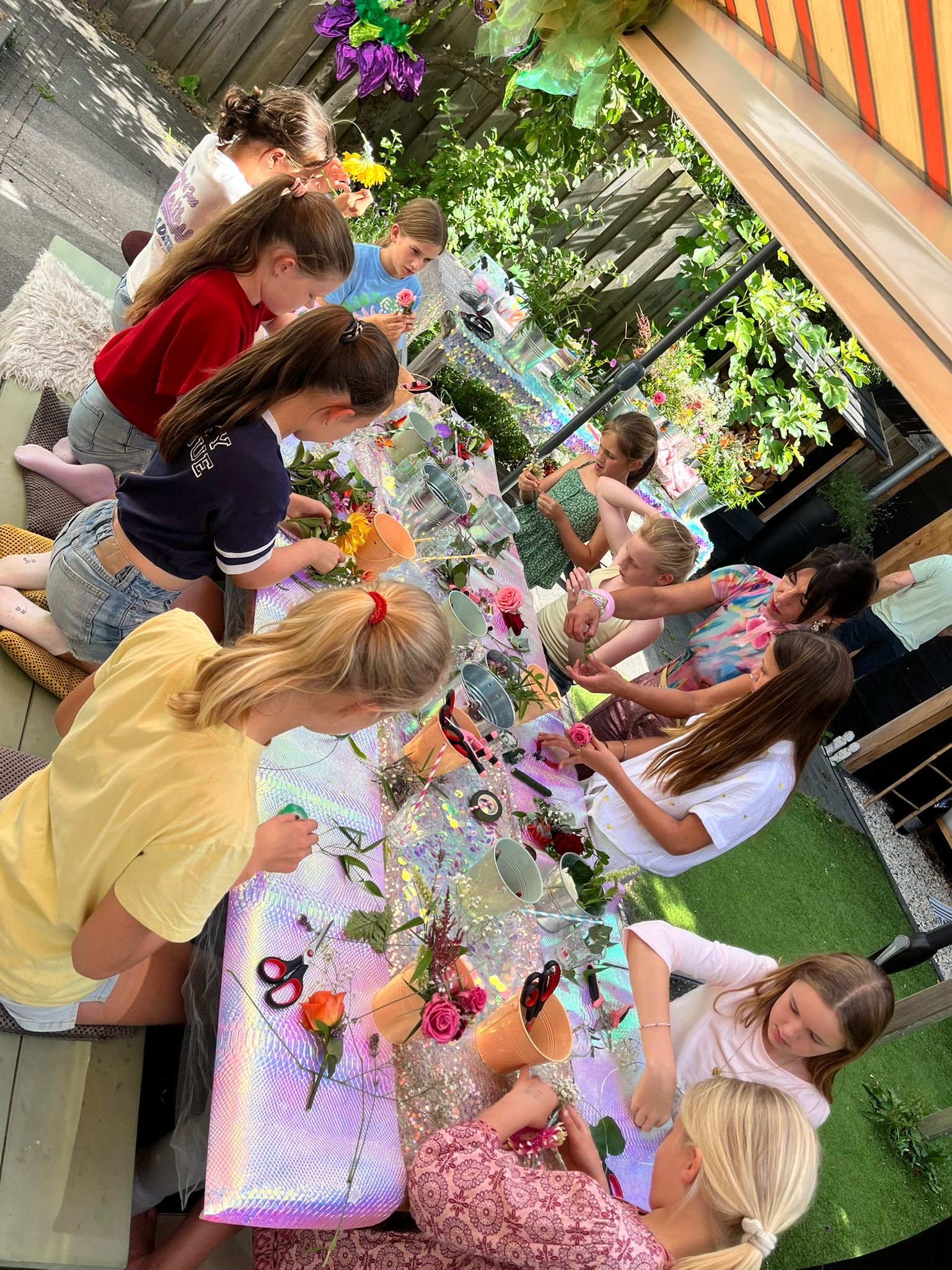 Children and adults gathered around a table outdoors, crafting flower arrangements with various flowers and tools, under a striped awning, surrounded by greenery and decorations.