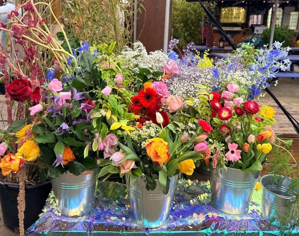 Colorful flower arrangements in silver buckets on a decorative table at an outdoor event.
