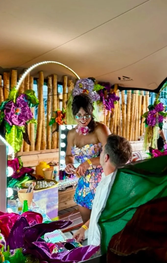 A woman dressed in a colorful, mosaic-style dress stands in front of a man seated in a chair. The woman is smiling and wearing glasses, with decorative flowers and greenery around her. They are in a decorated space with a mirror and lots of vibrant floral decorations.