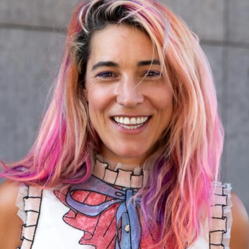A woman with pink and purple hair smiling outdoors in front of a gray wall, wearing a sleeveless top with a superhero graphic and a ruffled collar.