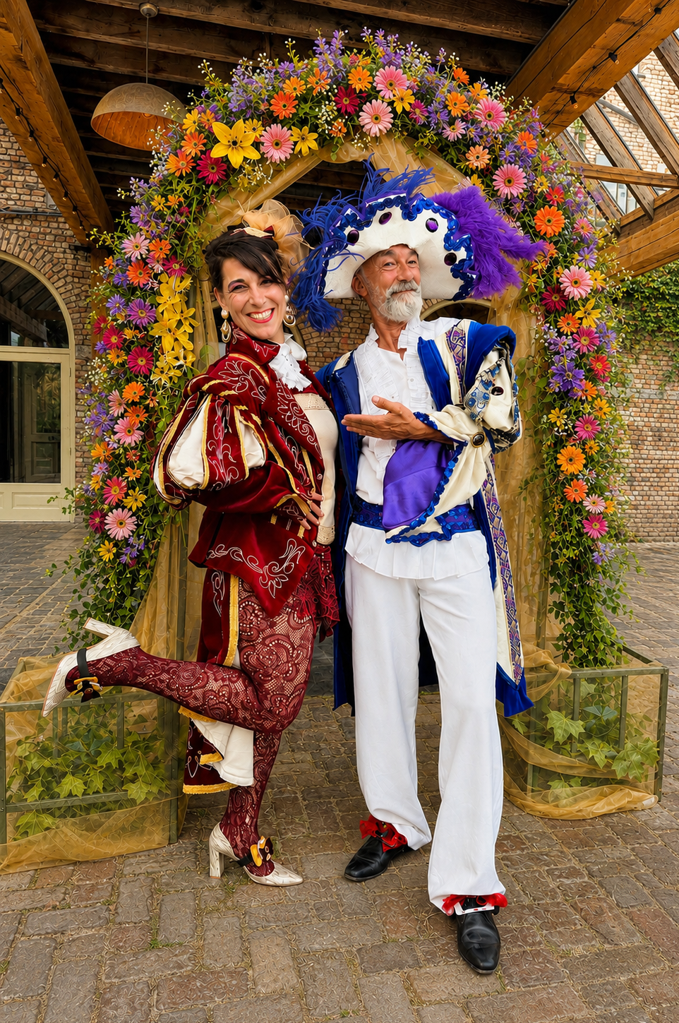 Two individuals dressed in colorful Renaissance costumes, standing under a floral archway, smiling at the camera.