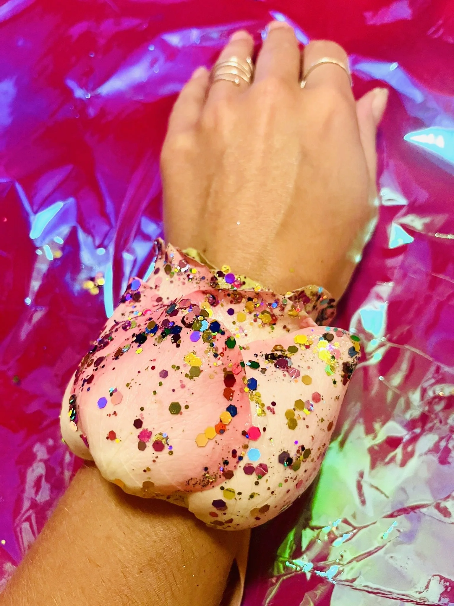 A hand with several gold rings rests on a pink donut covered in multicolored glitter sprinkles. The donut is inside shiny pink and green wrapping with reflective surfaces.