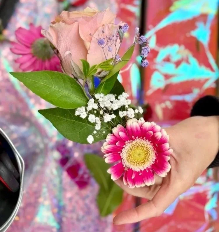 A hand holding a small bouquet of flowers with pink, purple, white, and green foliage, against a colorful background.