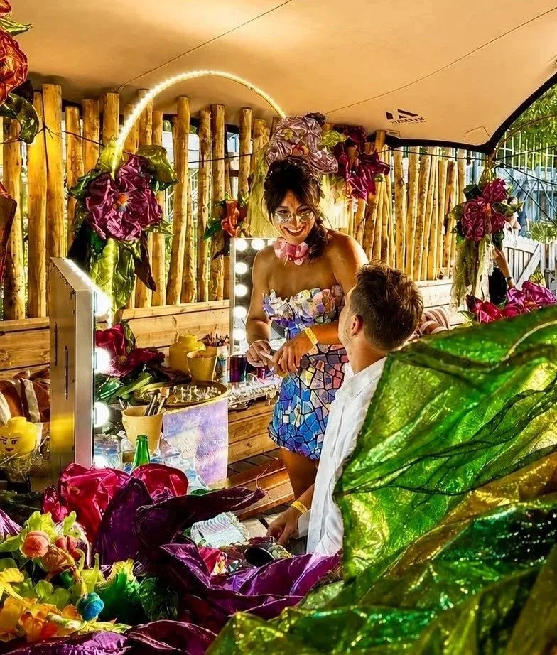 A woman with glasses and colorful outfit standing in front of a mirror, smiling at a man seated in front of her, surrounded by vibrant flowers and decorative plants in a tropical-themed setting.