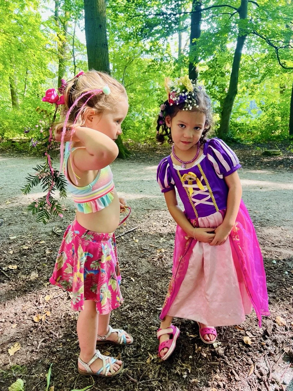 Two young girls standing in a forest with lush green trees in the background. One girl is dressed as a princess with a purple and pink dress, a floral crown, and pink sandals. The other girl is wearing a colorful striped crop top, a pink floral skirt, and sandals, with colorful beads and decorations in her hair.