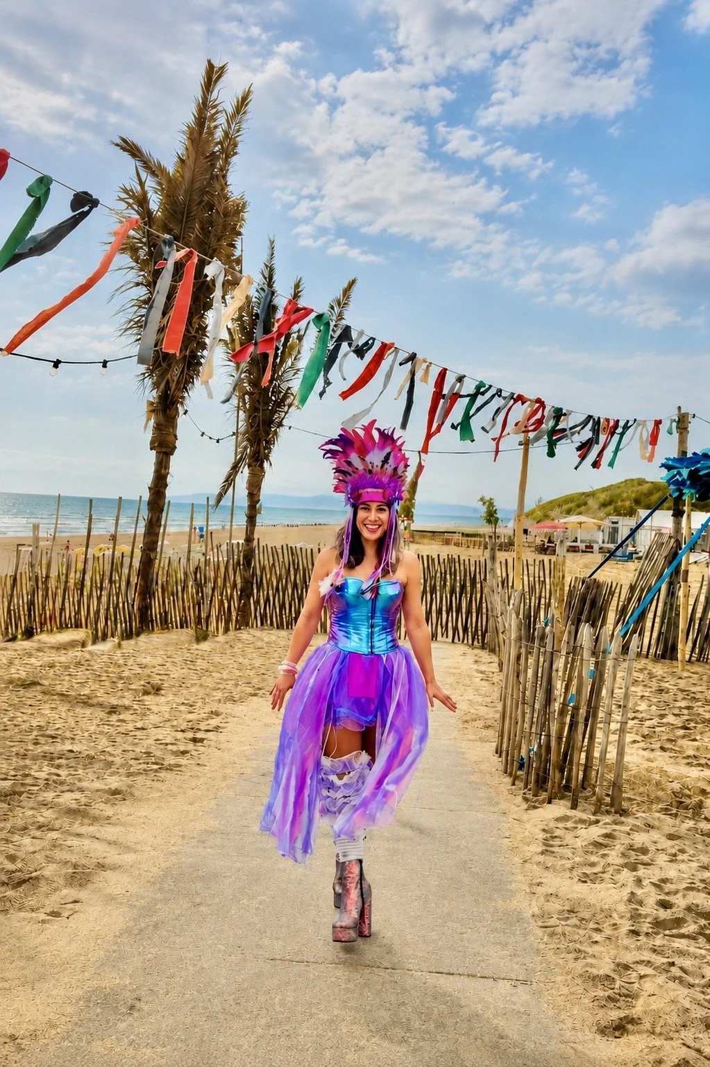 A women - Annelien de Bruin, performer, in a colorful costume with a feathered headdress walking on a sandy path at the beach, with palm trees, a decorated string of flags, and the ocean in the background.
