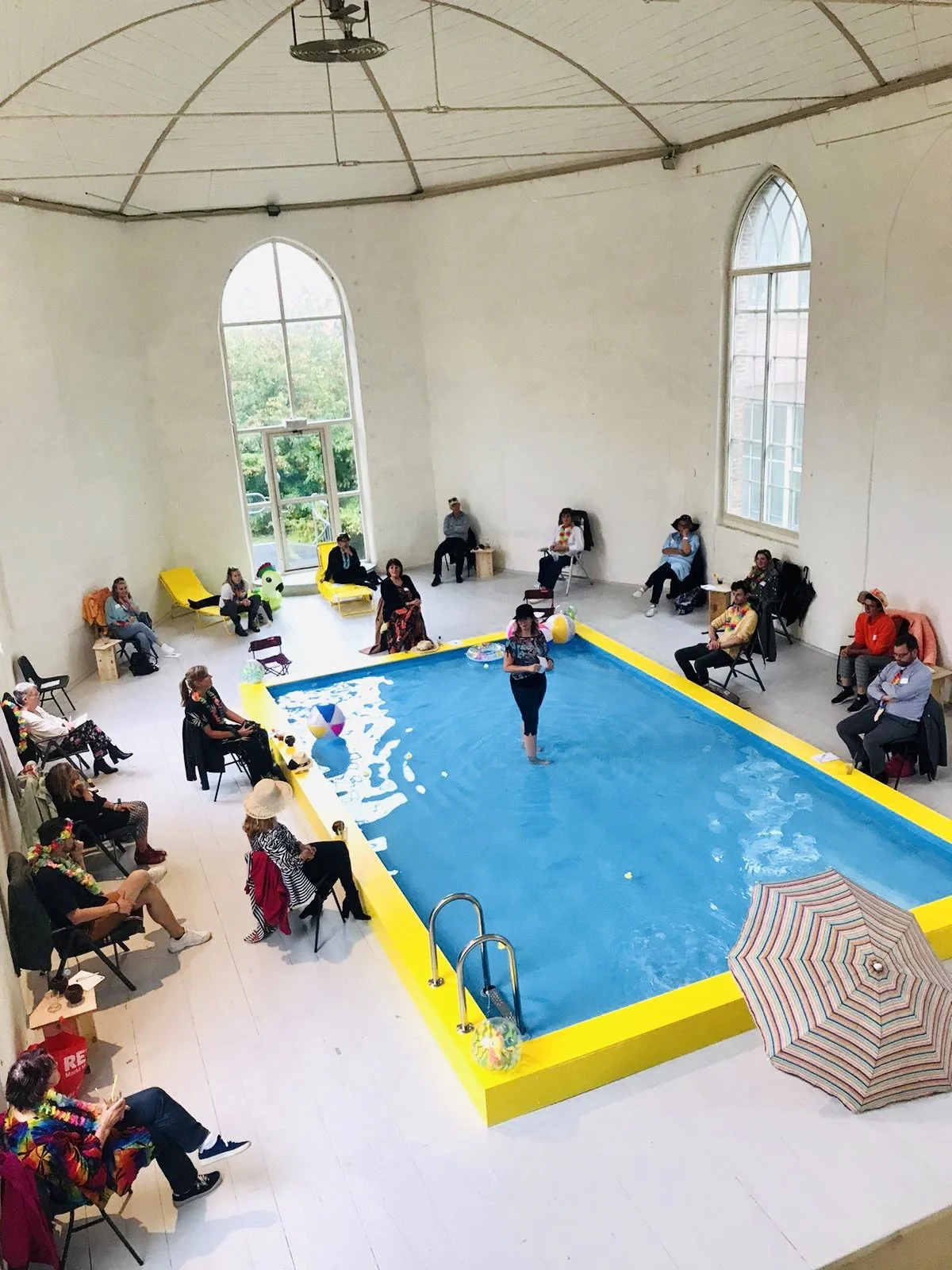 Indoor pool party in a spacious room with high arched windows and white walls, surrounded by seated guests in colorful attire, some wearing leis, with beach balls and floaties in the pool, a woman standing in the water, and an umbrella on the floor.