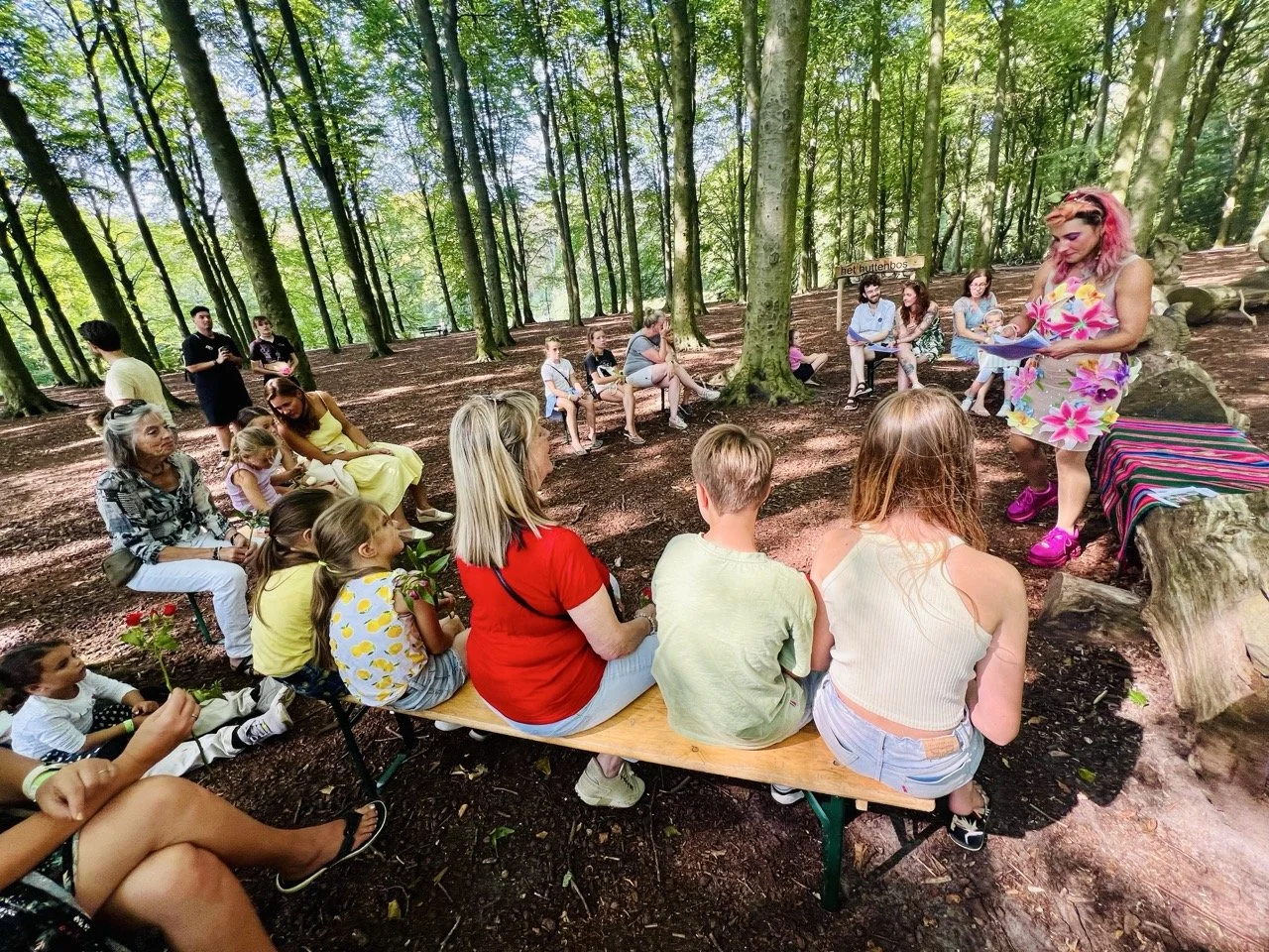 A group of children and adults gathered in a forest, listening to a woman with pink hair and a floral dress reading or speaking to them, seated on benches and logs surrounded by trees and shaded by leaves.