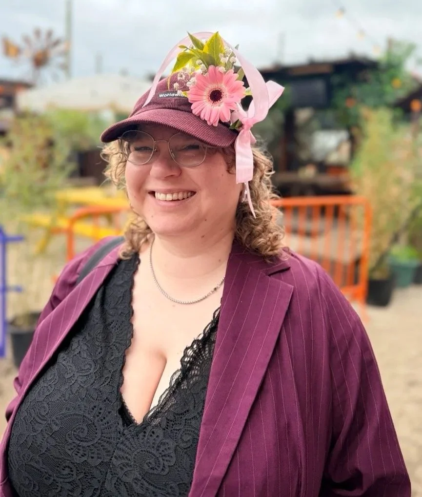 Woman smiling outdoors, wearing glasses, a maroon pinstripe blazer, black lace top, and a maroon cap decorated with a pink flower, greenery, and pink ribbon.