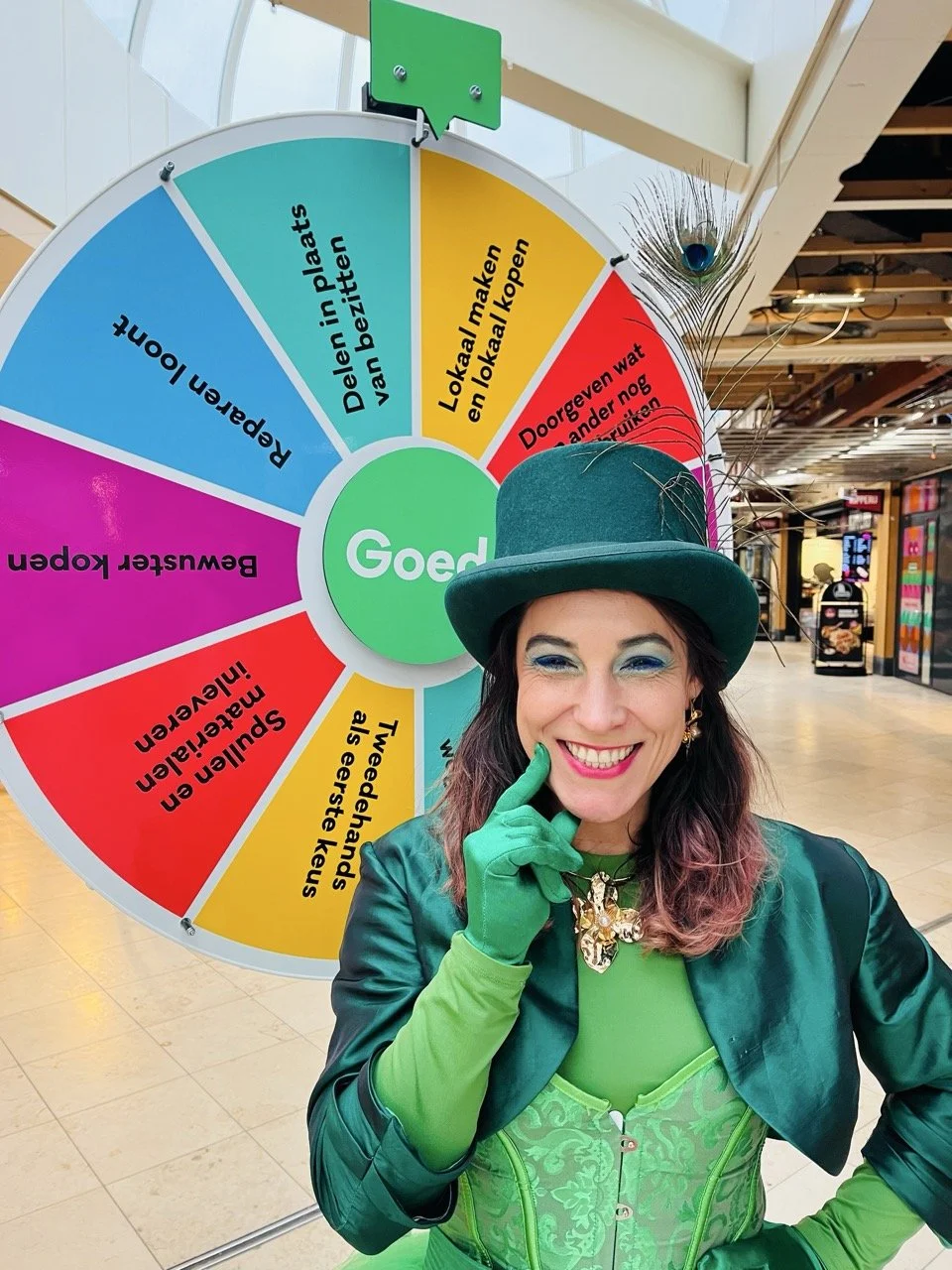 A woman dressed in a green costume with a green top hat and peacock feather, smiling and pointing to her cheek, standing in front of a colorful spinning wheel with Dutch text.