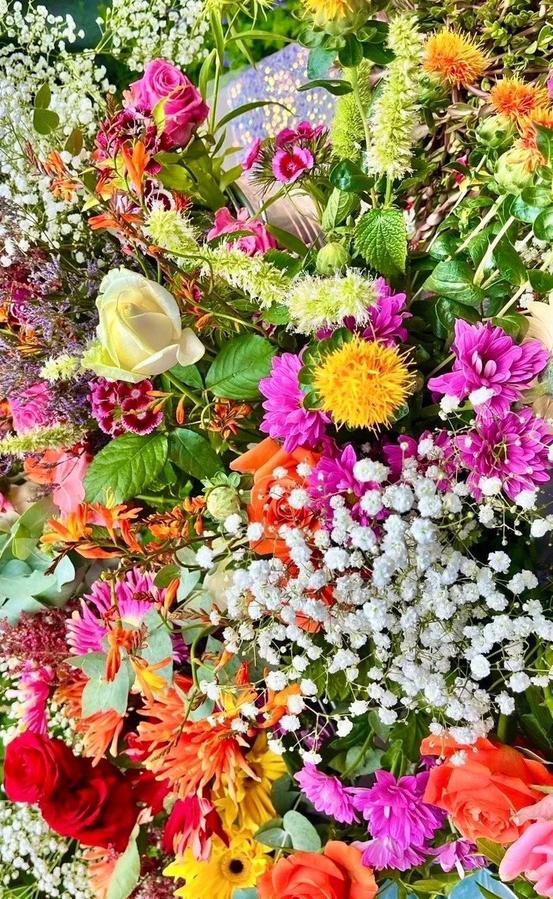 A vibrant bouquet of various colorful flowers including roses, daisies, baby's breath, and other mixed flowers with green leaves.
