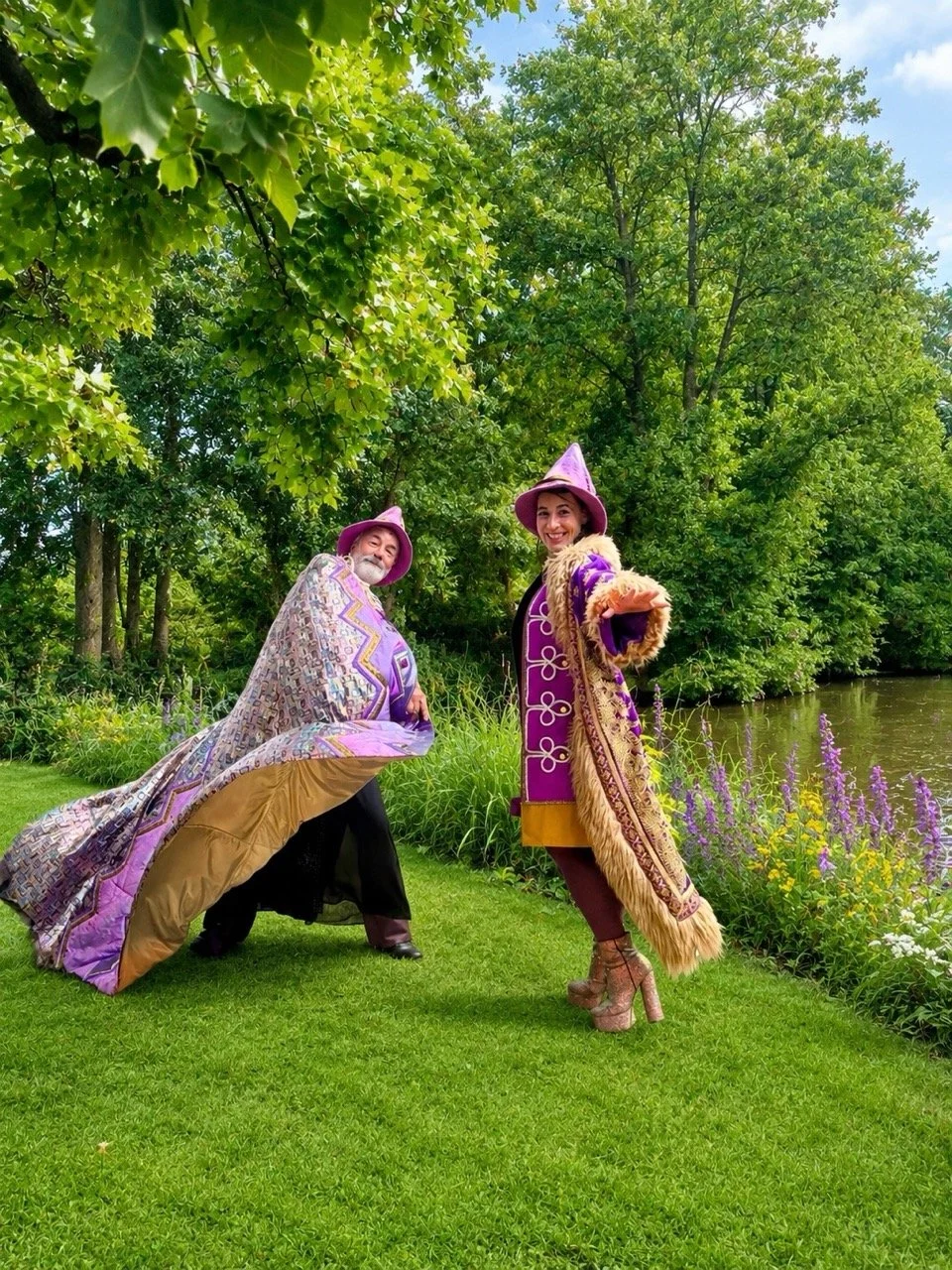 Two people dressed as wizards or magicians, wearing purple hats and robes with elaborate designs, standing on a grassy area by a small river or lake, surrounded by green trees and colorful flowers.