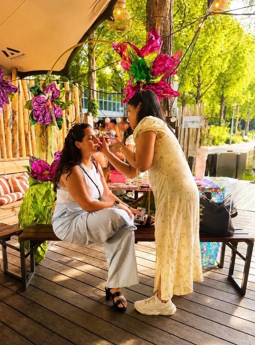 Woman sitting on a wooden bench getting makeup applied by another woman standing in front of her at an outdoor setting with green trees, lanterns, and colorful floral decorations.