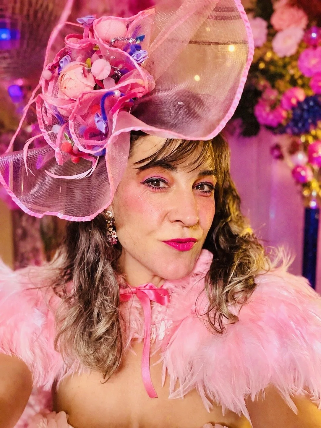 A woman dressed in pink with a large, decorative pink hat adorned with feathers, flowers, and ribbons, smiling in a colorful, floral-lit setting.