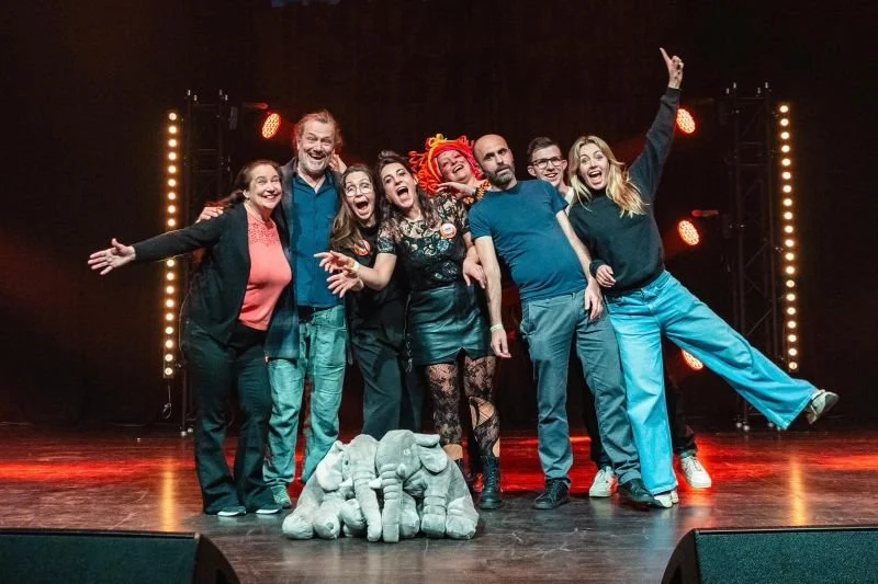 Group of seven people on stage, smiling and posing together with children and an elephant puppet, stage lights in background.