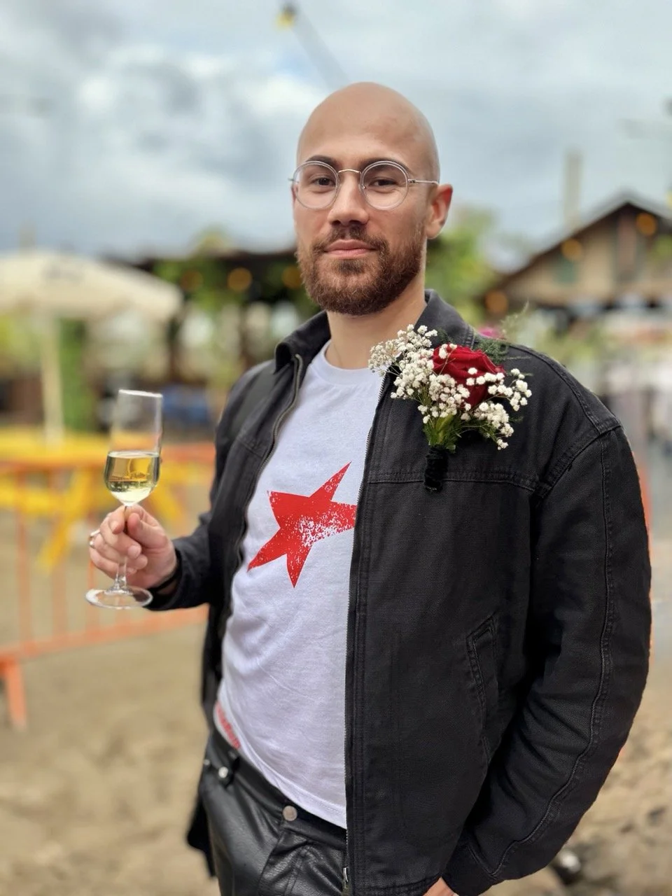 A man with a beard and glasses wearing a black jacket and white t-shirt with a red star holding a glass of champagne at an outdoor event. He has a boutonniere with red and white flowers pinned to his jacket.