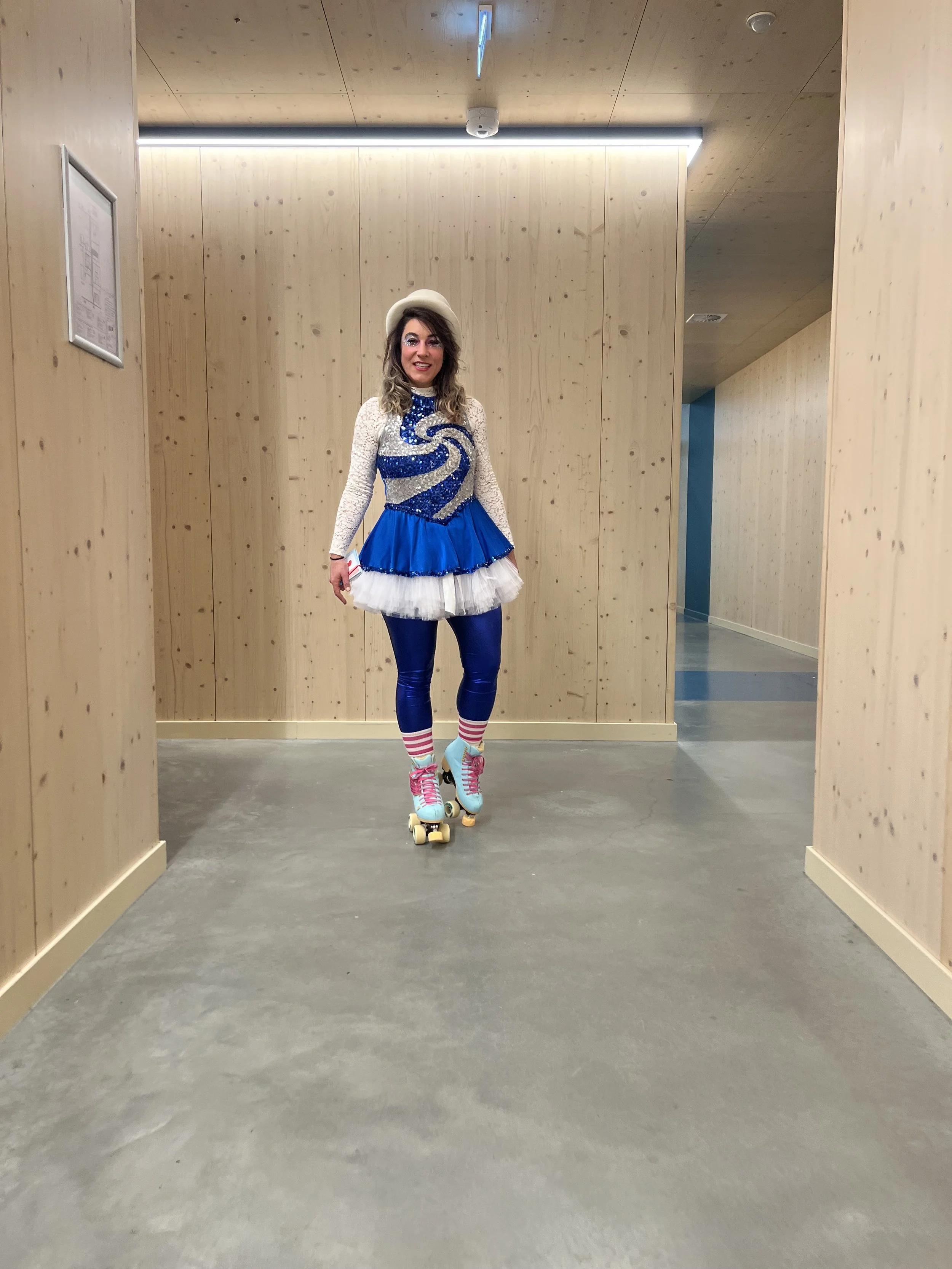 Woman in colorful roller skating outfit, with blue shiny leggings, striped socks, and colorful shoes, standing in an indoor corridor with wooden walls and ceiling.