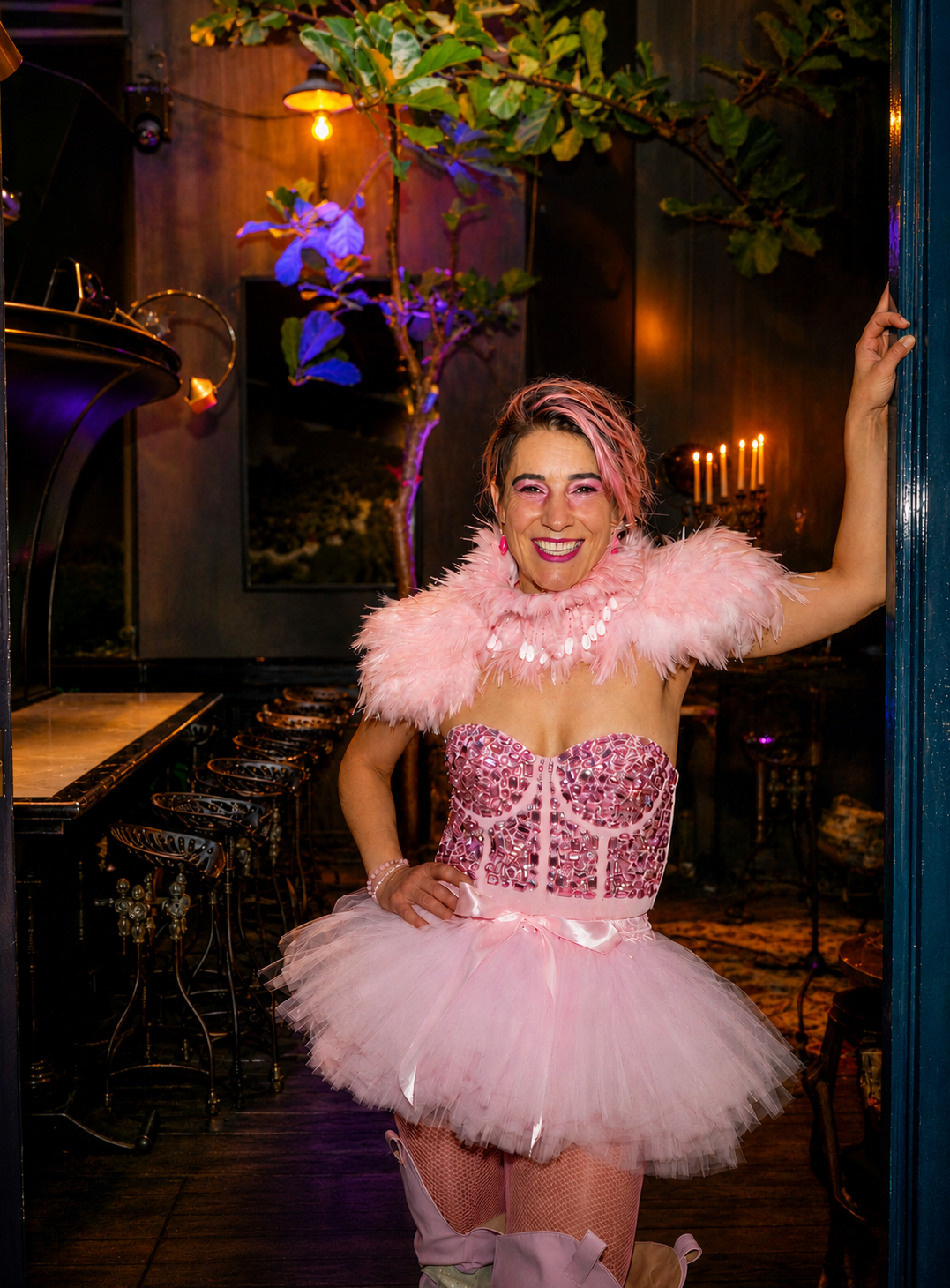 A woman dressed in a pink tutu and feathered shawl, smiling and posing inside a dimly lit room decorated with a tree, candles, and warm lighting.