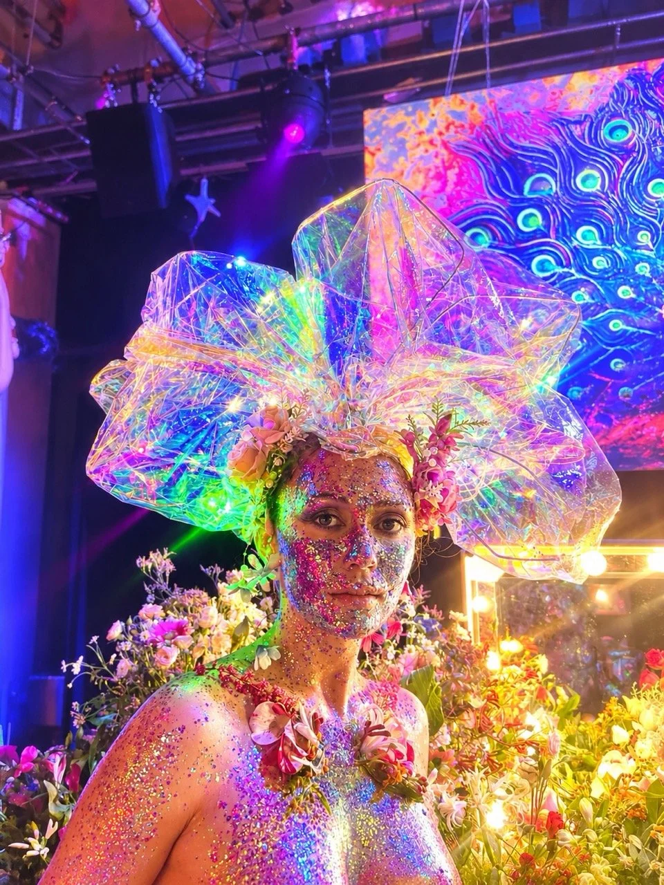 A person decorated with glitter and flowers, wearing a large, luminous, iridescent headpiece, surrounded by flowers and illuminated by colorful lights in a vibrant, artistic setting.