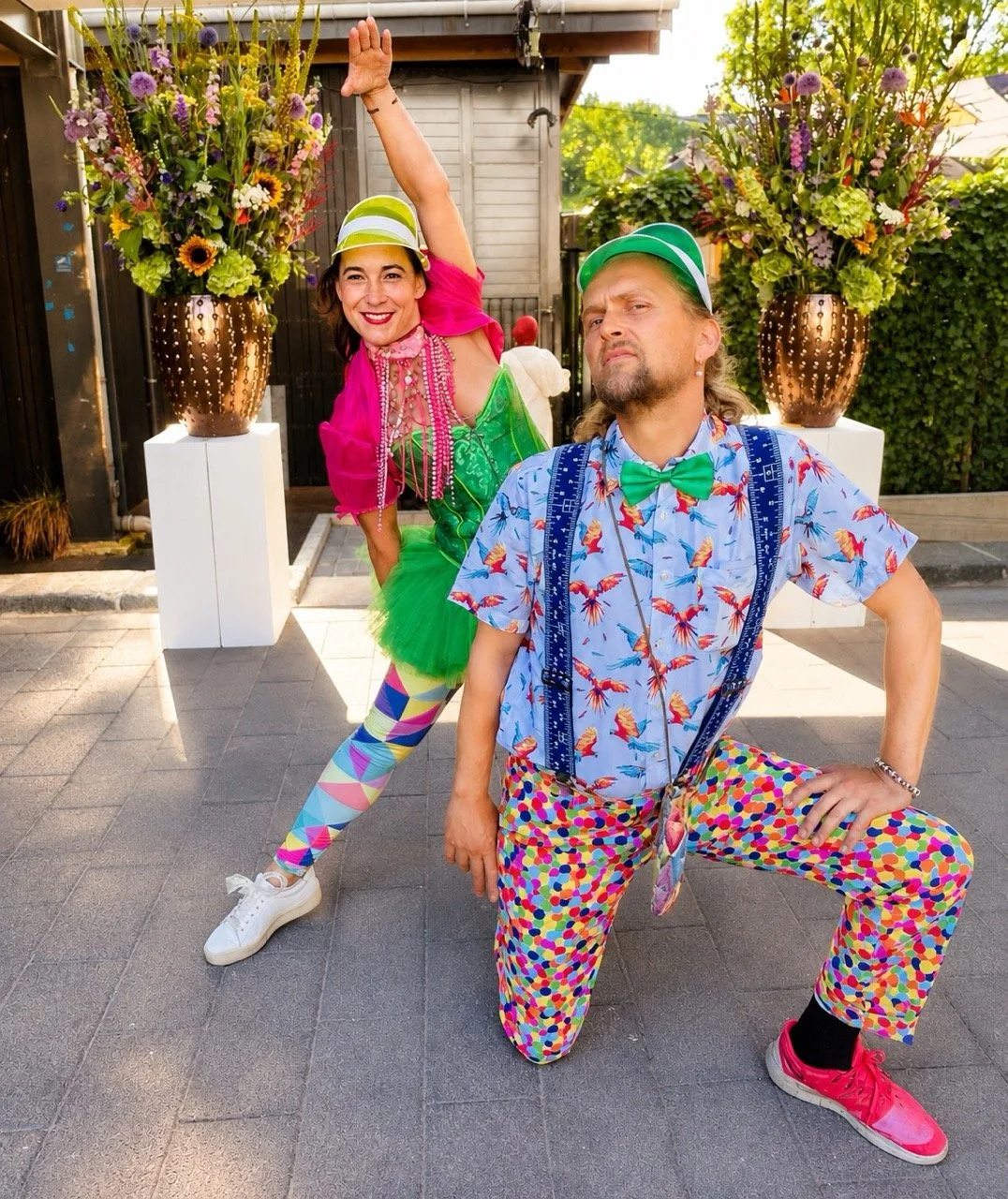 Two people dressed in colorful, playful costumes posing outdoors with large floral arrangements in copper vases behind them.