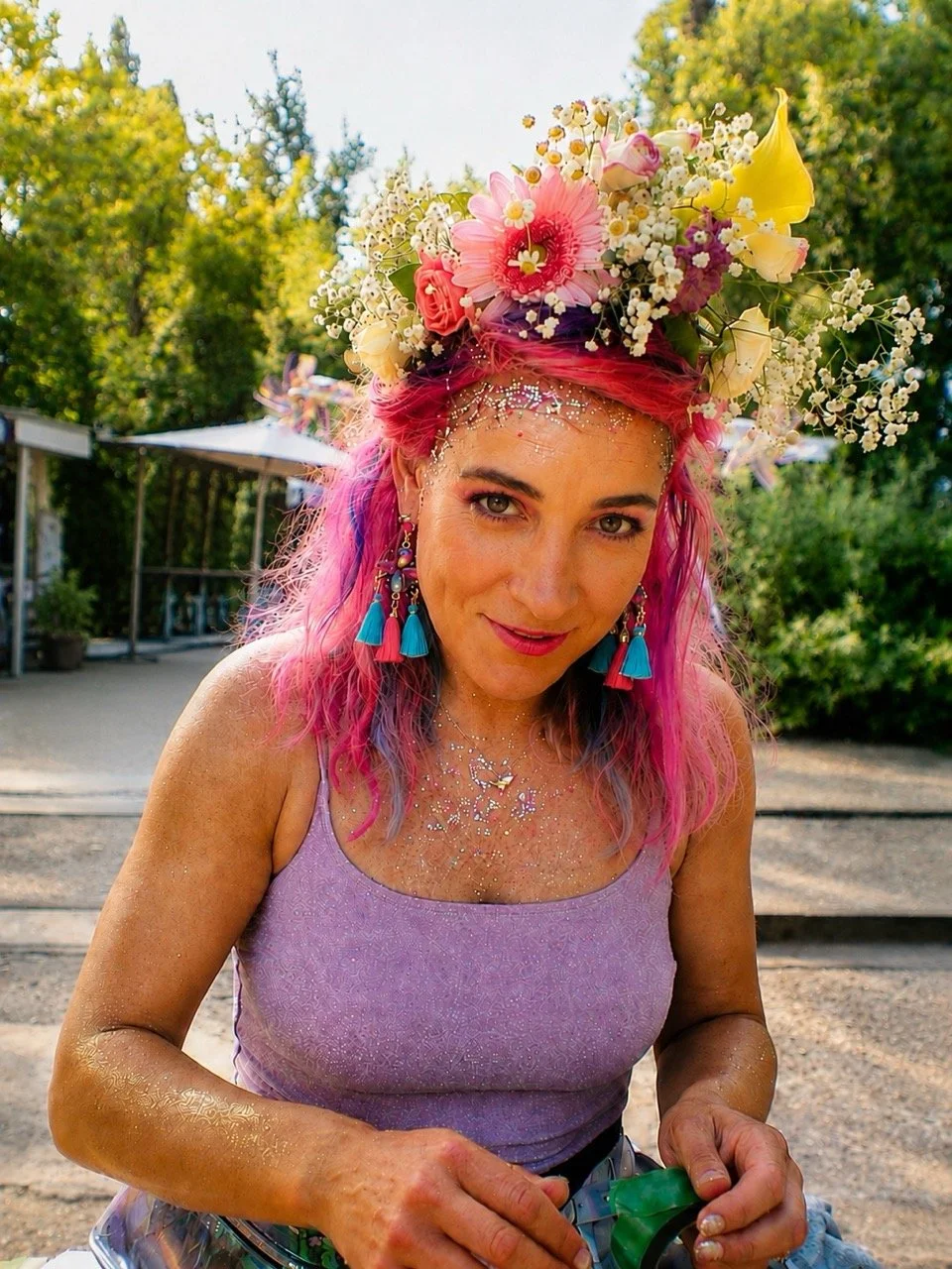 A woman with pink and purple hair wears a floral crown, glitter in her hair and face, and colorful earrings. She is outdoors with trees in the background, wearing a purple tank top.