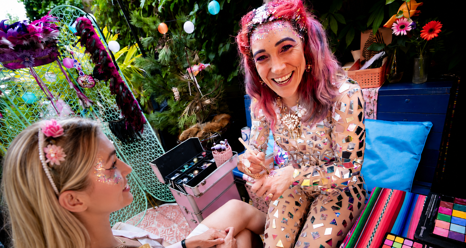 Two women with colorful makeup and glitter, one with bright pink hair, celebrating with makeup and accessories in a decorated outdoor setting featuring flowers, lights, and colorful decorations.
