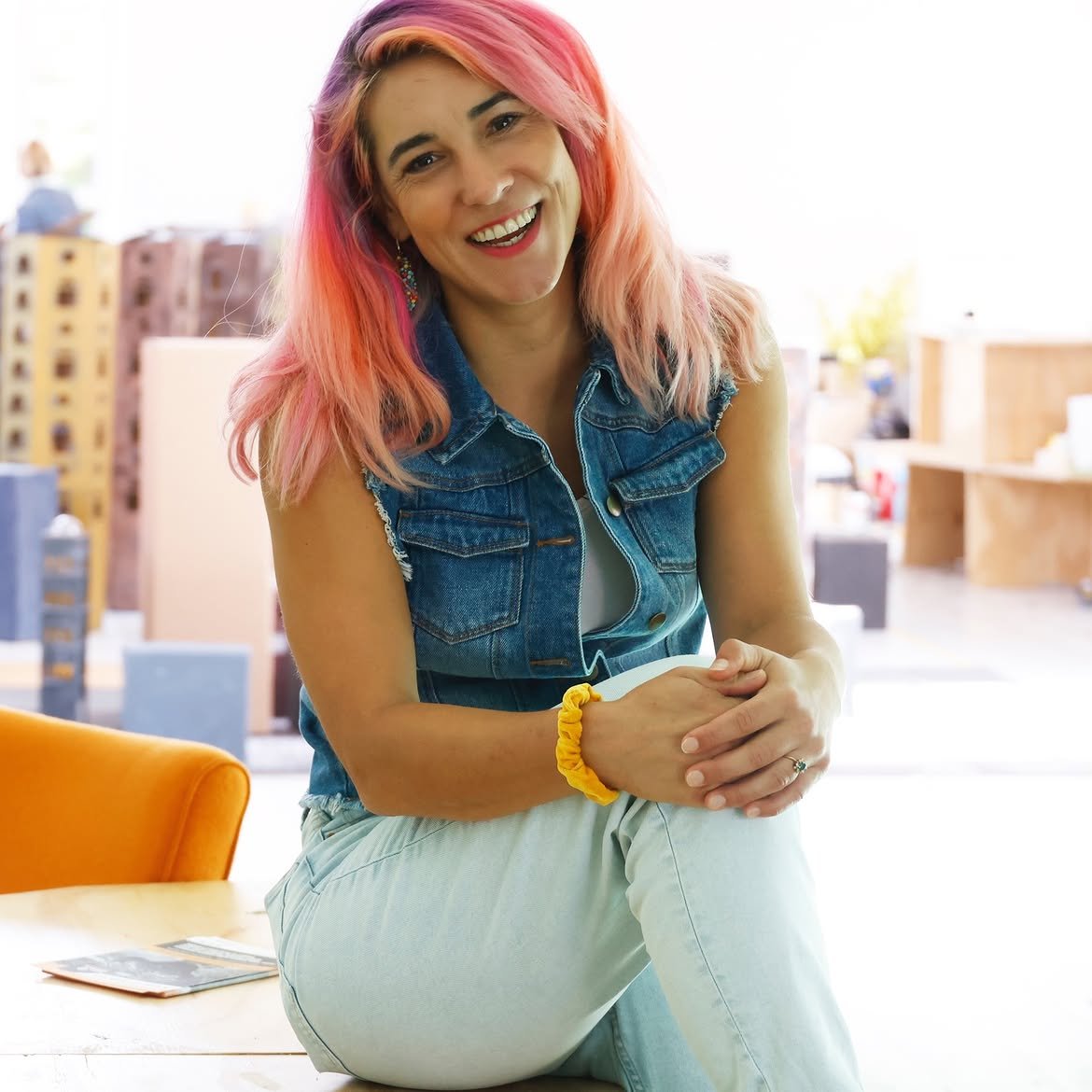 A woman with pink and orange hair smiling, sitting indoors with a bright background, wearing a denim vest and white pants.