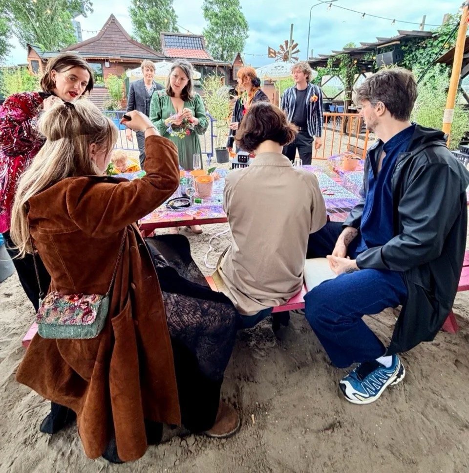 Group of people gathered outdoors around a pink picnic table, engaged in conversation, with some standing and others seated on benches, in a rustic setting with trees and wooden structures.